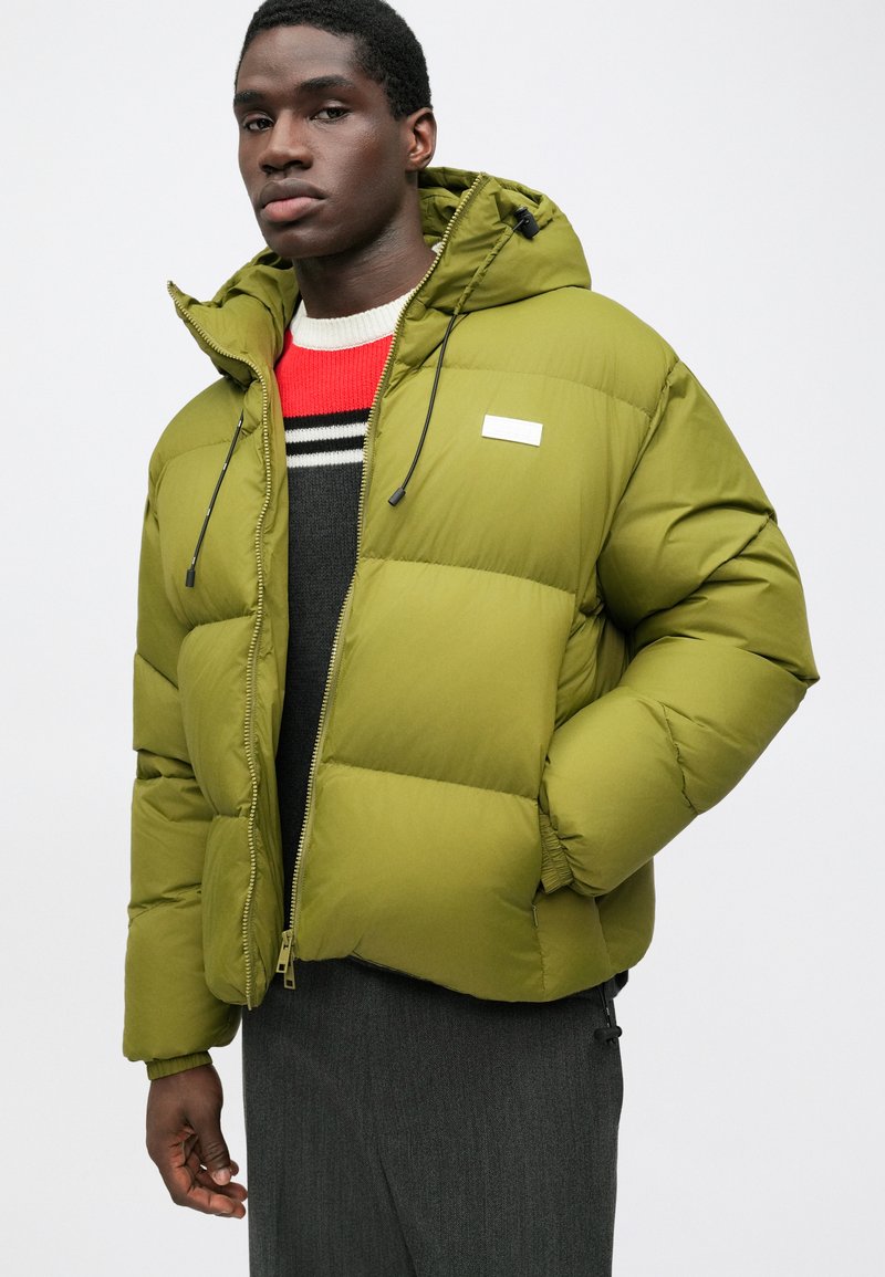Green puffer jacket with hood and adjustable drawstrings, featuring a front zipper and a small white logo on the chest. Textured, padded design.