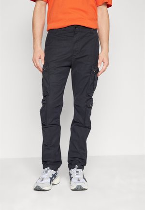 Mann trägt schwarze Cargo-Hosen mit mehreren Taschen, weiße und schwarze Sneakers und ein oranges Kurzarmhemd, steht vor einem schlichten Hintergrund.