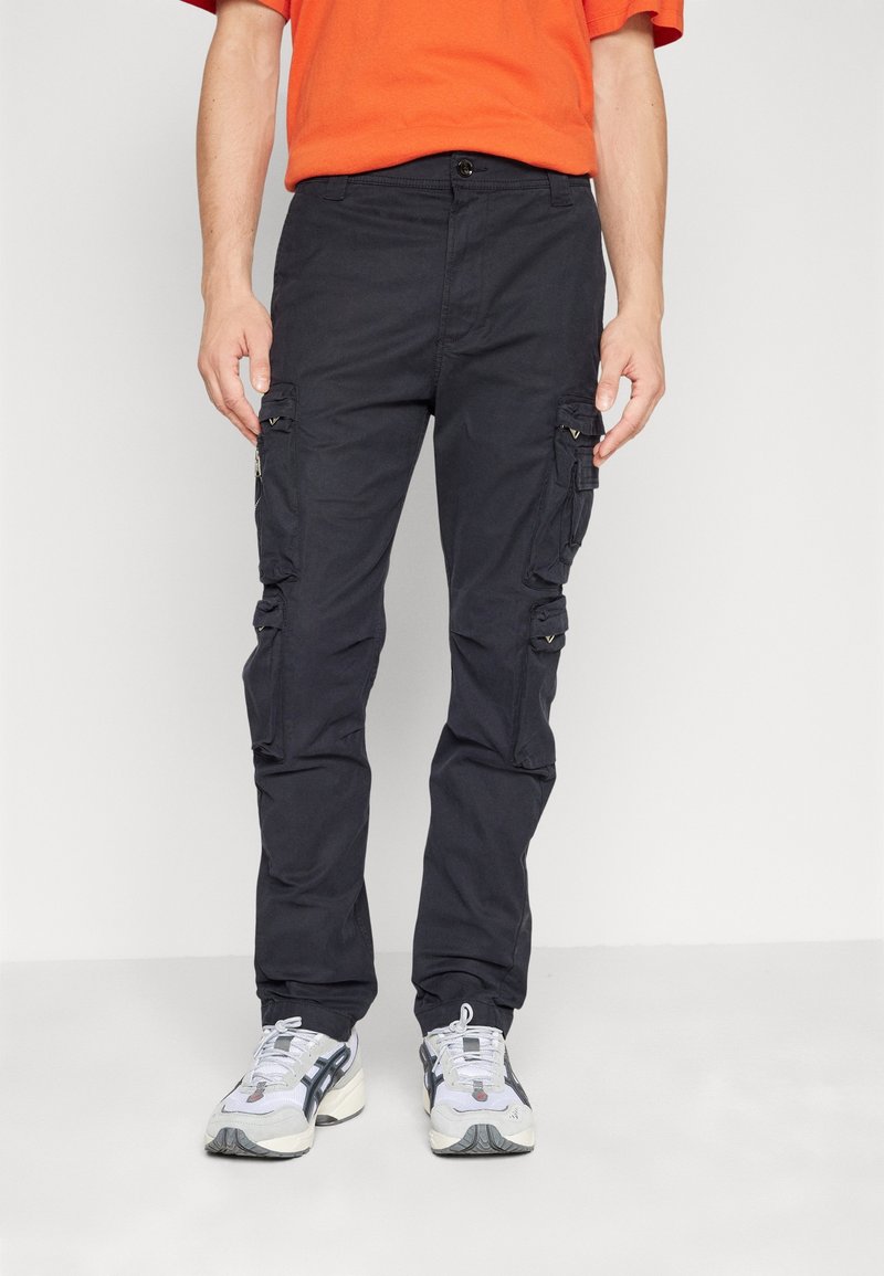 Homme portant un pantalon cargo noir avec plusieurs poches, des baskets blanches et noires, et un t-shirt orange à manches courtes, debout devant un fond uni.