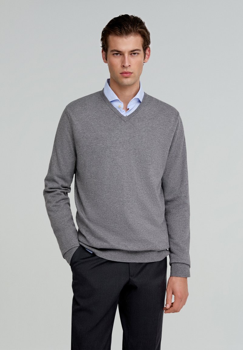 Suéter gris de cuello en v, hecho de un tejido suave y texturizado, combinado con una camisa de cuello azul claro. El modelo lleva pantalones oscuros y ajustados.