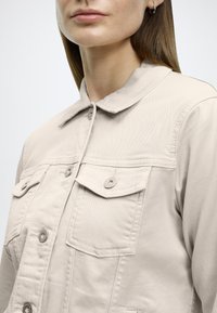 Ελαφριά μπεζ ζακέτα από απαλό ύφασμα, με κλασικό γιακά, κλείσιμο με κουμπιά, δύο τσέπες στο στήθος και διακριτικές ραφές.