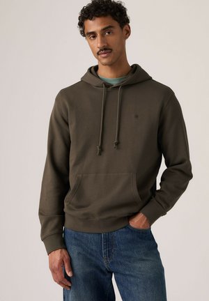 Homme aux cheveux bouclés et à la moustache portant un sweat à capuche vert olive foncé et un jean bleu, debout avec une main dans la poche.