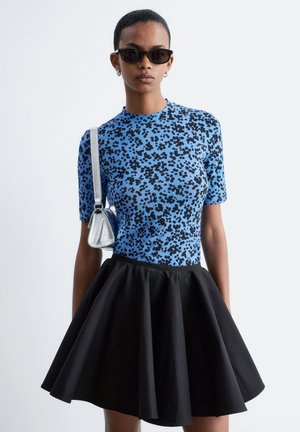 Person wearing black sunglasses, blue leopard print top, black flared skirt, and silver shoulder bag against plain background.