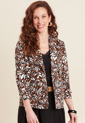 Blazer à motifs en crème, noir et rouille avec motif floral, fermeture à un bouton et détails de poches zippées. Cheveux bouclés et boucles d'oreilles créoles.