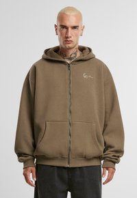 Haut zippé marron en tissu doux, avec une capuche à cordon, des poches avant et un petit logo blanc sur la poitrine.