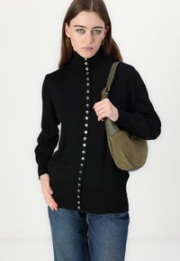 Magrone nero a collo alto con bottoni argento lungo la parte anteriore, abbinato a una borsa in suede verde e jeans a vita alta blu.