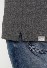 Grå bomullströja med en sidoventil vid nederkanten, med en liten vit logotag från The North Face på vänster sida.