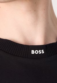 Chemise noire avec un col côtelé. Le col présente un texte blanc au dos qui indique "BOSS" centré. Texture de tissu lisse.