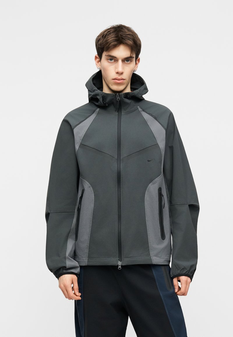 Junger Mann trägt eine dunkelgraue Kapuzenjacke mit hellgrauen Einsätzen und schwarzen Reißverschlusstaschen, steht vor einem einfarbigen Hintergrund.