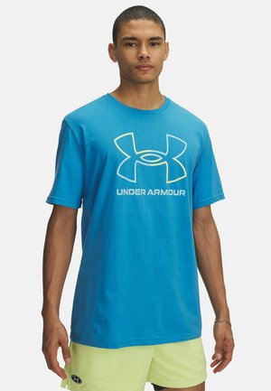 Camiseta de algodón azul con logo en perfil amarillo y manga corta. Cuello redondo y corte recto, llevada sobre pantalones cortos de malla verde claro.