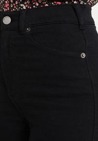 Jeans de mezclilla negra con botón y remaches plateados, usados con una blusa de patrón floral metida por dentro.