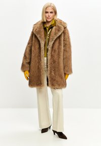 Vaaleatukkainen nainen, jolla on yllään suuri ruskea karvatakki, keltaiset käsineet, oliivinvihreä takki, kermaiset housut ja ruskeat korkokengät, seisomassa valkoisella taustalla.