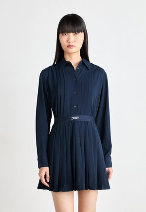 Abito plissettato navy con scollo a collare, maniche lunghe, chiusura con bottoni e una cintura con marchio. Tessuto liscio e lunghezza a metà coscia.