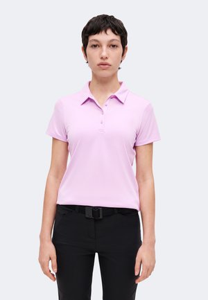 Ung person med kort mørkt hår iført en lyserød kortærmet polo-shirt og sorte bukser med sort bælte, stående foran en ensfarvet baggrund.