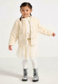 Cream faux fur jacket with heart-shaped pockets, layered over a lace dress. Sheer skirt, white tights, and silver lace-up boots complete the outfit.