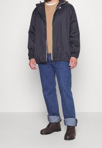 Giacca leggera nera con cappuccio, dettaglio della zip gialla, abbinata a un maglione beige, jeans blu e stivali marroni con lacci.