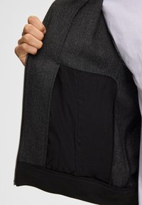 Giacca nera con chiusura a zip sul fronte, caratterizzata da un esterno texturizzato e una fodera interna nera. Un grande taschino visibile all'interno.
