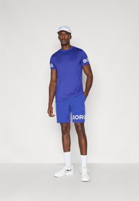 Μπλε αθλητικό μπλουζάκι και σορτς με το "BORG" τυπωμένο σε άσπρο. Φορεμένο με άσπρο καπέλο και άσπρες κάλτσες, συνδυασμένο με λευκά αθλητικά παπούτσια.