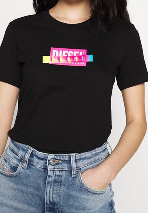 Kvinde iført sort kortærmet skjorte med farverigt Diesel-logo og lyseblå højtaljede jeans med hånden i lommen.