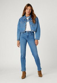 Chaqueta de denim recortada de color azul claro con dos bolsillos frontales, combinada con unos vaqueros azules de tiro alto. La modelo lleva una camiseta blanca debajo.