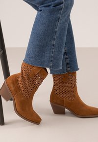 Botines de ante marrón con detalles trenzados en la caña, punta cuadrada y tacón bajo. Combinados con pantalones de mezclilla azules.