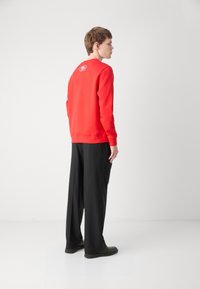 Sweatshirt vermelho com um logótipo redondo nas costas, emparelhado com calças largas pretas e calçado escuro, apresentado contra um fundo branco liso.