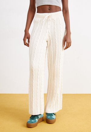 Woman wearing cream cable-knit wide-leg pants and teal platform sneakers, standing on a yellow floor against a plain background.