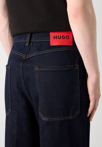 Nærbillede af bagsiden af mørkeblå jeans med orange syninger og et rødt label med teksten "HUGO" på livet, båret med en sort top.