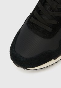 Fekete sportcipő sima szintetikus felsőrésszel és texturált velúr ráhordásokkal. Fekete fűzővel és kis tüskékkel ellátott fehér gumitalppal rendelkezik.