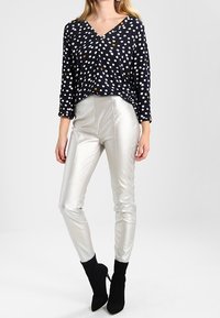 Marineblaues Oberteil mit weißen und orangefarbenen Punkten, V-Ausschnitt, Dreiviertelärmel; metallic glänzende silberne enge Hose; schwarze Stiefeletten.