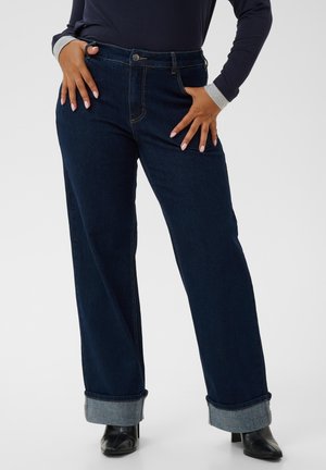 Personne portant un jean bleu foncé retroussé, des bottes noires à talons et un haut noir à manches longues avec des poignets gris, les mains posées sur les hanches.