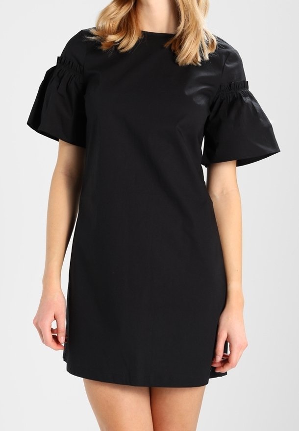 Femme portant une robe courte noire avec des manches bouffantes et des détails froncés sur le haut des bras, debout devant un fond clair uni.