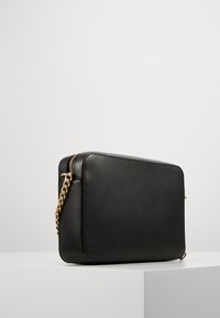 Svart rektangulär axelväska i läder med guldkedjestrap och dragkedja, placerad på en vit piedestal mot en enfärgad bakgrund.