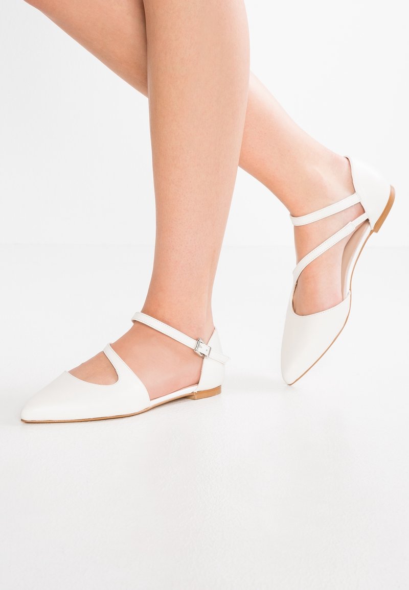 Jambes portant des chaussures plates blanches à bouts pointus avec double bride à la cheville sur fond blanc.