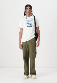 Tricou alb din bumbac cu un imprimeu grafic colorat, asociat cu pantaloni cargo verzi măslin, dotati cu buzunare. Modelul poartă adidași bej.