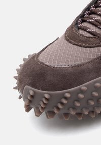 Smešane smeđe tenisice s teksturiranim gornjim dijelom od tkanine uz anekse od antilopa, gumeni potplat s izraženim šiljcima i složenim detaljima šivanja.