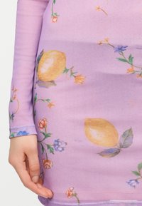 Ένα λιλά φόρεμα με μακριά μανίκια που έχει εκτύπωση με λεμόνι και πολύχρωμα λουλούδια, με ένα χέρι να ακουμπάει στο πλάι.