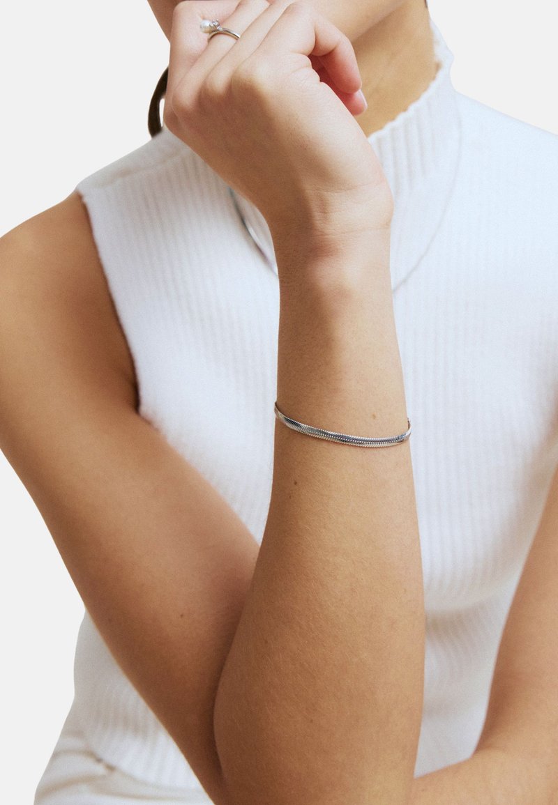 Armband van zilverkleurig metaal met een glad, gestroomlijnd ontwerp. Wordt gedragen om een pols, met een geribbelde witte top op de achtergrond. Simpel en minimalistisch.
