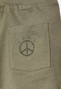 Calções de algodão verde-oliva com um bolso com remendo. Apresenta um símbolo da paz e o nome da marca impressos no bolso com tinta verde mais escura.