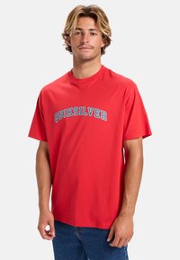 Camiseta de algodón roja con un logo azul de "QUIKSILVER" en el pecho. Manga corta, cuello redondo, corte relajado, textura suave.