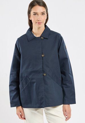 Femme portant une veste bleu marine boutonnée avec de grandes poches frontales et un col, associée à un pantalon blanc, debout devant un fond uni.