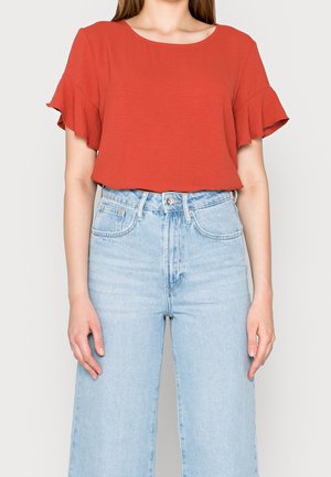 Mujer con una blusa naranja de manga corta con volantes, metida dentro de unos vaqueros de mezclilla azul claro de talle alto, sobre un fondo liso.