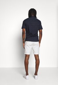 Camicetta navy a maniche corte con motivo a pois bianchi, abbinata a pantaloni corti bianchi arrotolati. Il modello è visto da dietro su uno sfondo semplice.