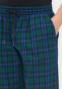 Pantalons à carreaux verts et noirs avec des accents bleus, taille élastique et cordon de serrage. Comprend deux poches latérales et une texture douce.