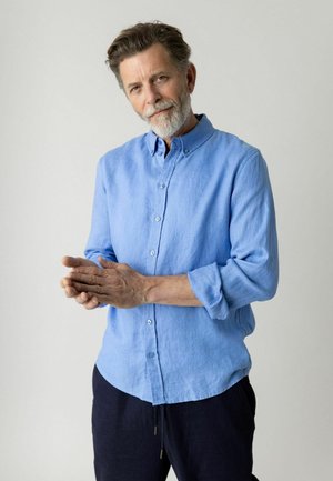 Vidutinio amžiaus vyras su pilka barzda, dėvintis šviesiai mėlyną mygtukais užsegamą marškinių ir tamsias kelnes, stovintis priešais paprastą šviesų foną.