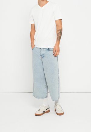 Homme portant un T-shirt blanc à col en V, un jean large bleu clair raccourci, des chaussettes blanches et des baskets blanches avec des semelles marron, debout les mains dans les poches.