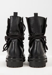 Par de botas de combate de cuero negro con suelas gruesas, cordones enrollados alrededor de los tobillos, vistas desde atrás sobre una superficie blanca.