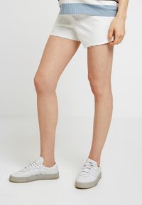 Vita denimshorts med fransar i nederkant, kombinerat med vita sneakers med en texturerad sula. Toppen har ett ribbat mönster i blått och vitt.