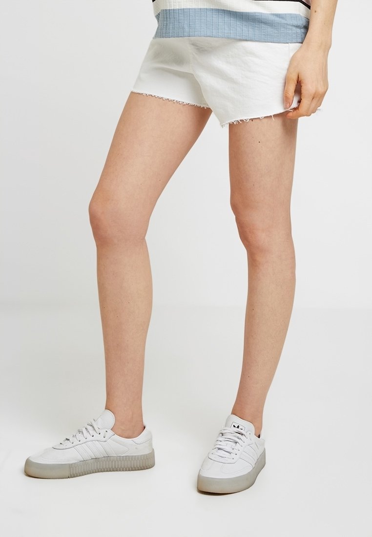 Vita denimshorts med fransar i nederkant, kombinerat med vita sneakers med en texturerad sula. Toppen har ett ribbat mönster i blått och vitt.