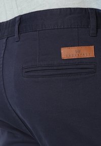 Navy blue pants with a smooth texture, featuring a back pocket and a rectangular leather tag with "Kronstadt" embossed on it.